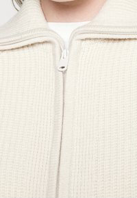 Kermansävyinen, teksturoidulla pinnalla varustettu vetoketjullinen takki, jossa on korkea kaulus ja vaakasuora ribbineuloskuvio, valkoinen vetoketju ja tyylikäs muotoilu.