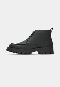 Bottine noire en cuir avec semelle épaisse nervurée, lacets à l’avant et languette au talon, vue de profil sur fond clair.