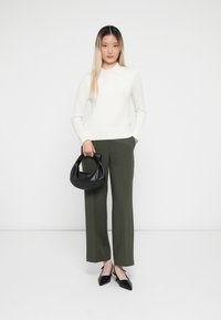 Bílé pletené polo svetr, olivové široké kalhoty a černá kožená kabelka. Modelka má na sobě špičaté černé podpatky. Jednoduchý, moderní design.