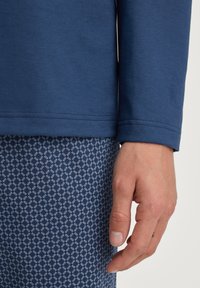 Marineblauwe lange mouw top met een gestructureerde afwerking en omgeslagen mouwen, gecombineerd met een patroonbroek met kleine witte diamantvormen op een donkere achtergrond.