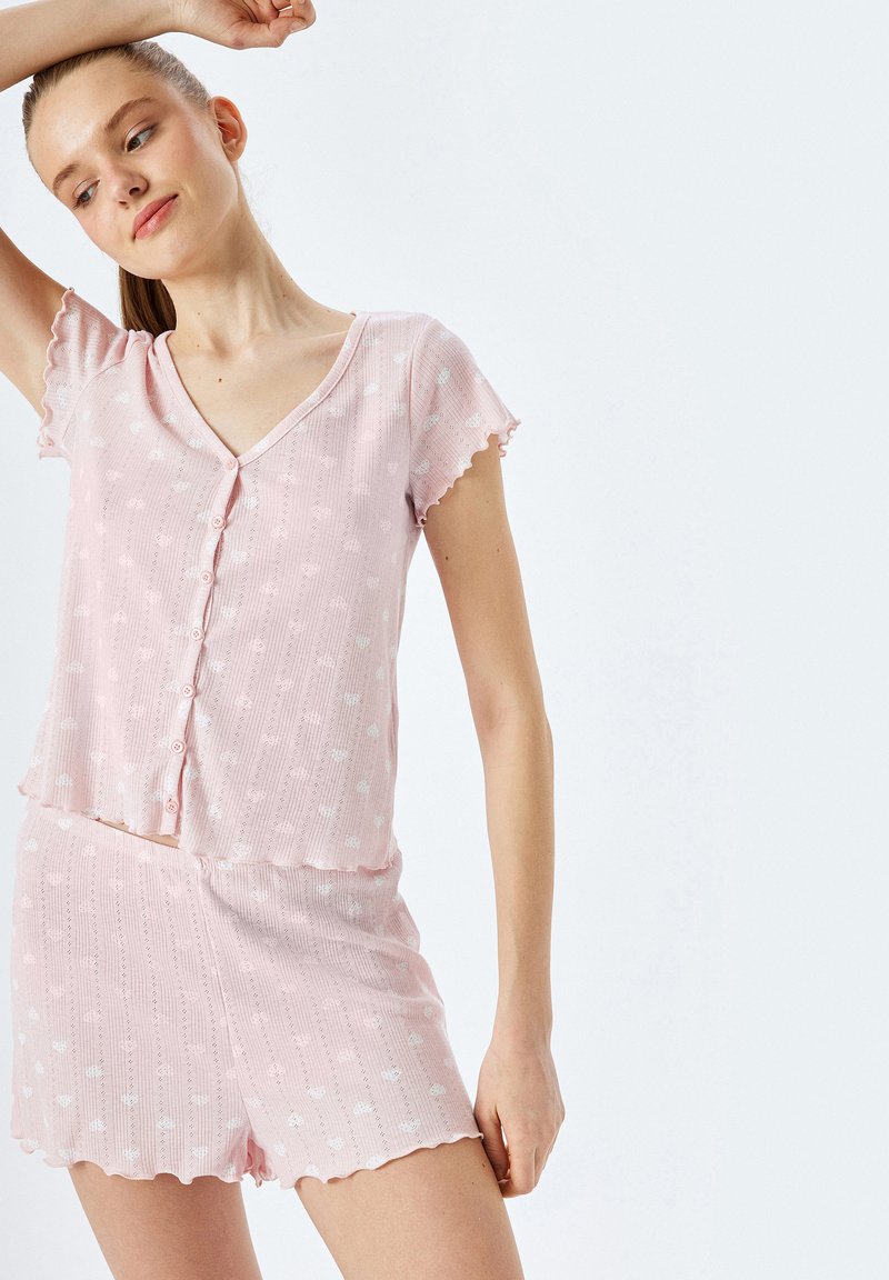 El conjunto de pijama de manga corta en color rosa cuenta con una parte superior de cuello en V con detalles de botones y bordes ondulados, acompañado de pantalones cortos a juego cubiertos con un patrón sutil.