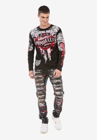 Schwarzes Langarmshirt mit weißen und roten grafischen Designs, kombiniert mit ausgebeulten schwarzen Jeans mit mehrfarbigen Farbklecksen. Weiße Sneaker.