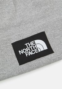 Berretto lavorato a maglia grigio con etichetta nera che presenta il logo bianco di "The North Face", mostrando un design semplice e un tessuto testurizzato.