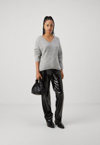 Grau geripptes Sweatshirt mit V-Ausschnitt, kombiniert mit glänzenden schwarzen Kunstlederhosen. Schwarze Tasche mit gerafften Griffen. Deutlich ausgeprägte Texturen.