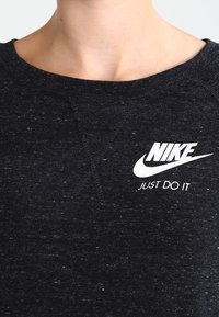 Detailní záběr na černou melírovanou mikinu s bílým logem Nike a textem "Just Do It" na horní části hrudníku.