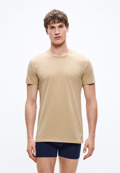 Μπεζ κοντομάνικο μπλουζάκι με στρογγυλή λαιμόκοψη, κατασκευασμένο από λεία ύφασμα, με λεπτομέρεια διακριτικού λογότυπου κοντά στο τελείωμα, φορεμένο πάνω από σκούρα μπλε αντρικά μποξεράκια.