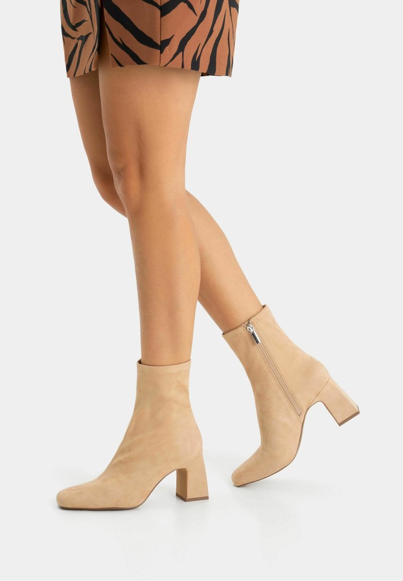 Jambes portant des bottines en daim fauve à talons carrés et fermetures éclair latérales, associées à une jupe à motifs marron et noir.