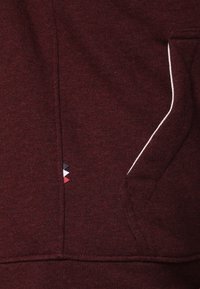 Burgundy tröja med en strukturerad yta, med en vit detalj längs fickan och en liten tri-färg logotyp.