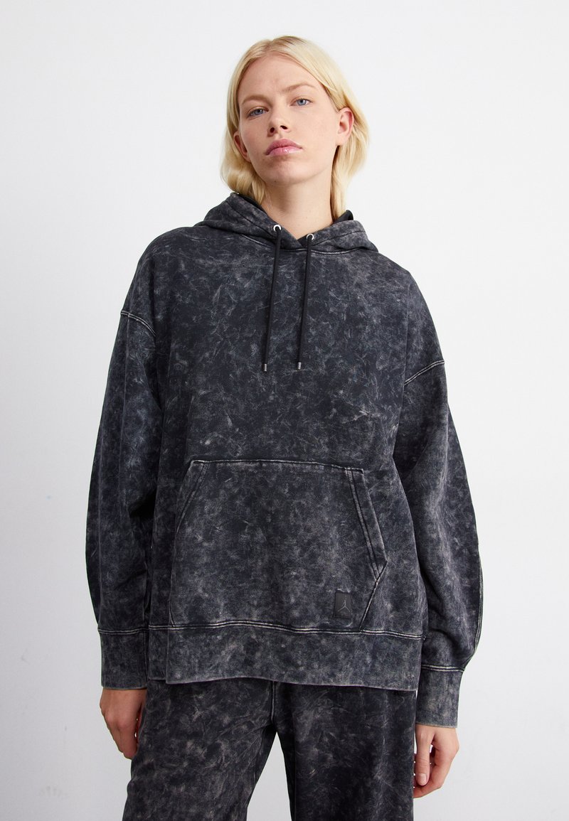 Sudadera negra de gran tamaño en tejido desgastado, con bolsillo frontal tipo canguro, capucha con cordón y un sutil acento de logo. Textura suave.