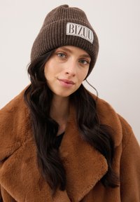 Vrouw met lang donker haar, een gebreide bruine muts met het label "BIZUU" en een grote bruine bontjas van nep, tegen een effen lichte achtergrond.
