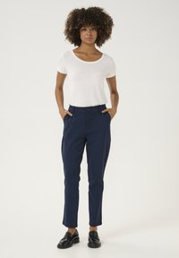 Pantalons ajustés bleu marine avec un devant plat, des poches latérales et une fermeture à bouton, associés à un t-shirt blanc ajusté et des mocassins noirs.
