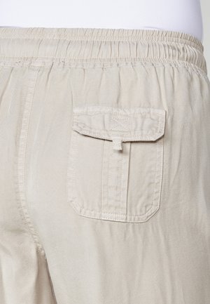 Le pantalon cargo beige présente une poche arrière avec un rabat, une taille élastique et un tissu texturé, mettant en avant un design pratique et décontracté.