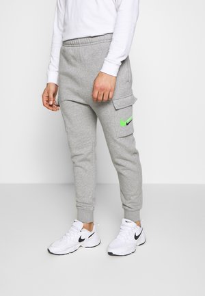 Mann trägt graue Nike-Sweatpants mit grünem und schwarzem Logo an der Seitentasche und weiße Nike-Sneaker und steht vor schlichter Hintergrund.