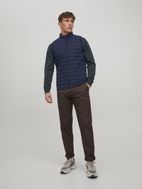 Gilet matelassé navy avec col haut, associé à une chemise à manches longues vert foncé et un pantalon marron ; baskets beiges avec des accents gris.