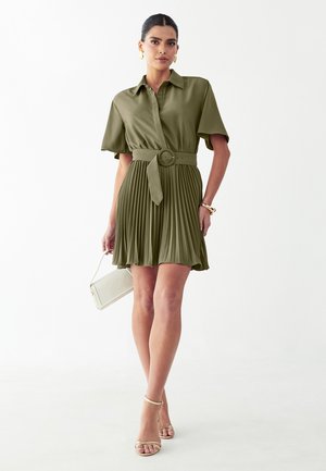 Mujer con un vestido corto de manga corta color verde oliva con cinturón y falda plisada, sosteniendo un bolso blanco, pendientes de aro dorados y tacones beige.