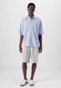 Camisa de manga corta azul claro con rayas verticales, bolsillo a la izquierda, combinada con pantalones cortos deportivos grises y zapatillas grises y blancas.