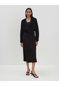 Cardigan noir en tricot avec une ouverture devant, associé à une robe noire ajustée. Le modèle porte des tongs noires à talons. Design simple et épuré.