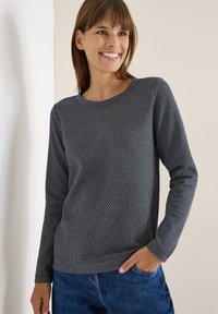 Marineblauer Pullover mit einem strukturierten geometrischen Muster, rundem Ausschnitt, langen Ärmeln und einer lockeren Passform. Kombiniert mit blauen Jeans.