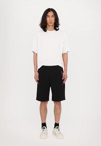 Pantaloni sportivi neri con tasche laterali, abbinati a una maglietta bianca oversize. Le scarpe sono sneakers bianche; lo sfondo è bianco uniforme.