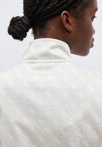 Personne aux cheveux tressés attachés en chignon porte une veste de couleur claire avec un motif et le logo "GUESS" sur le col arrière.