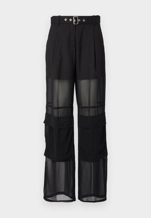 Schwarze Hose mit weitem Bein, einem Gürtel mit silberner Schnalle und geschichteten transparenten Paneelen im unteren Bereich.