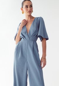 Modra kombinezona z globokim V-izrezom, kratkimi volumnimi rokavi in nabrano sprednjo stranjo. Narejena iz teksturirane tkanine, ima raven pas in široke hlačnice.