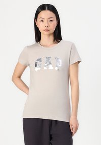 Beige kortermet t-skjorte laget av bomull, med et sølvreflekterende "GAP"-logo over brystet. Myk tekstur og rund hals.