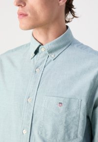 Camicia button-up azzurro chiaro con colletto, taschino sul petto e texture sottile. Presenta un piccolo logo ricamato sulla tasca.