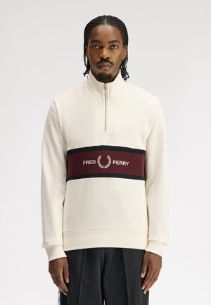 Mann mit geflochtenem Haar, der einen cremefarbenen Pullover mit einem kastanienbraunen und schwarzen Streifen sowie dem "Fred Perry"-Logo auf der Brust trägt und vor weißem Hintergrund steht.