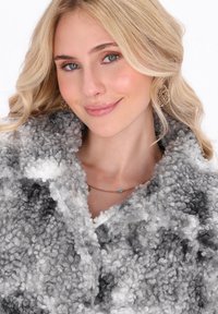 Mujer rubia con el cabello rizado, que lleva pendientes de filigrana dorados, un collar de cuentas turquesas y un abrigo de lana gris y blanco con textura.