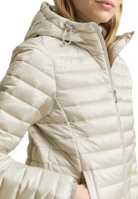 Lichtgewicht, roomkleurige pufferjack met horizontale doorgestikte structuur, een capuchon met trekkoorden en een voorste ritssluiting.