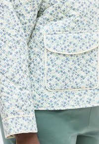 Κοντινό πλάνο μανικιού και τσέπης από σακάκι με ανοιχτόχρωμο λουλουδένιο σχέδιο, φορεμένο με πράσινο παντελόνι σε άτομο με σκούρο δέρμα.