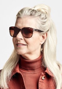 Kameleonttiväriset aurinkolasit ruskeilla linsseillä, joissa on pyöreä muoto ja sulava muotoilu. Yhdistetty syksyisin rustinväriseen poolokaulukseen ja takkiin.