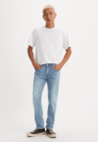 Tânărul cu păr scurt blond stă cu mâinile în buzunare, purtând un tricou alb simplu, blugi albaștri deschiși, adidași negri și cercei din argint.