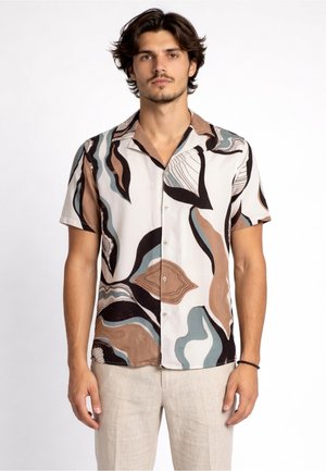 Giovane uomo con capelli scuri che indossa una camicia a maniche corte con bottoni, decorata con motivi astratti marroni, neri e blu, e pantaloni di colore chiaro.