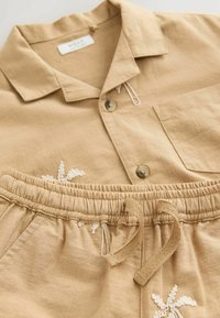 Camicia di lino color sabbia con colletto e tasca frontale, decorata con dettagli ricamati bianchi; pantaloni corti abbinati con vita elasticizzata ecordino.