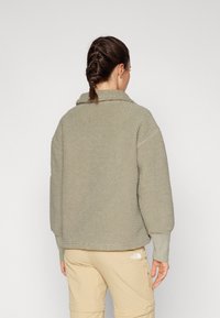 Giacca in pile verde oliva morbido con colletto, spalle cadenti e polsini a coste, abbinata a pantaloni beige con tasca laterale con zip.