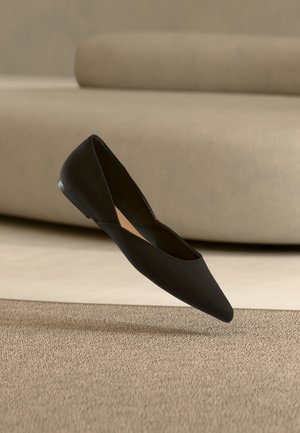 Μαύρο, επίπεδο παπούτσι με μυτερή μύτη κρεμασμένο πάνω από ένα μπεζ υφασμάτινο χαλί με έναν καναπέ σε ουδέτερο χρώμα στο φόντο.