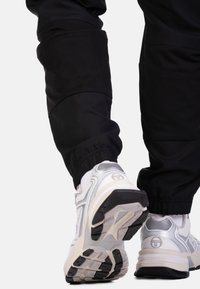 Zwarte sportieve joggers met manchetten bij de enkels, in combinatie met witte en zilveren sneakers met textuurranden en subtiele merkemblemen.
