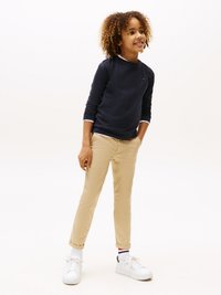 Navy long-sleeve shirt with white trim, tan slim-fit pants, and white sneakers with blue and red accents. Curly hair, smiling expression.