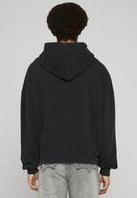 Melnā kapučjakas džemperis ar brīvu piegriezumu, nolaistām pleciem un ribām aprocēs. Raksturo mīkstu tekstūru un vienkāršu dizainu.