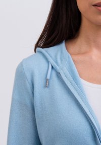 Ljusblå hoodie i mjukt tyg med dragsko i huvan och silverdetaljer samt dragkedjestängning, visad nära kragens område.