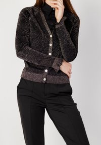 Cardigan noir et argenté pailleté avec un col en V, une fermeture à boutons et des poignets côtelés. Associé à un pantalon noir, doté de poches latérales.