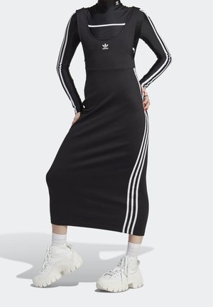Osoba nosząca czarną sukienkę Adidas z białymi paskami po bokach, w połączeniu z czarną bluzką z długim rękawem i białymi masywnymi sneakersami.