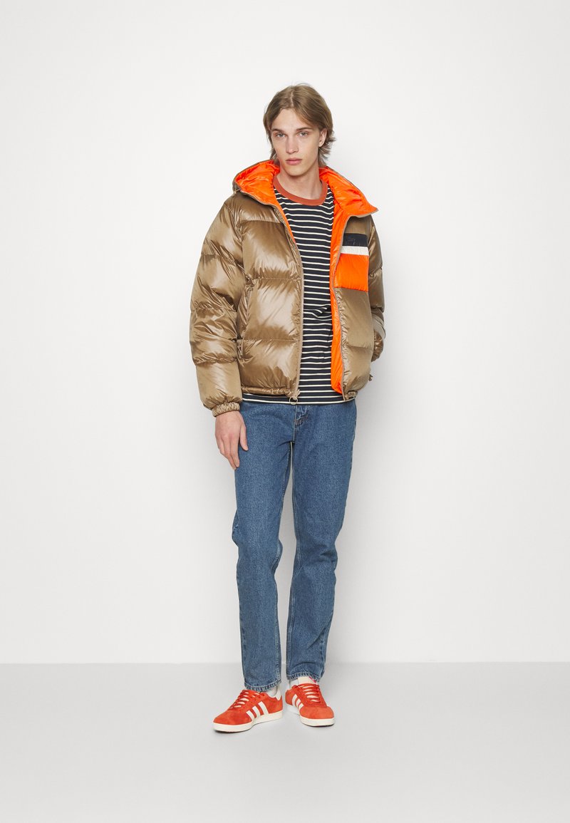 Puffjacke in glänzendem Braun mit einer orangefarbenen Kapuze und Brusttasche, kombiniert mit einem schwarz-weiß gestreiften Shirt und blauen Jeans.