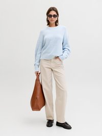 Vrouw draagt een lichtblauwe trui, beige broek, zwarte schoenen, donkere zonnebril, houdt een grote bruine leren tas vast, staat tegen een effen achtergrond.