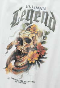 White T-shirt featuring a skull surrounded by flowers and a snake. Text above reads "ULTIMATE Legend," with shadows and vibrant colors.