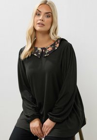 Donna bionda seduta, che indossa una maglietta nera a maniche lunghe con una texture a coste e un colletto a fiori su uno sfondo semplice.