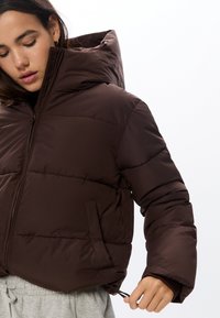 Brązowa kurtka puchowa z kapturem, z pikowanym wzorem, z zamkiem błyskawicznym z przodu i bocznymi kieszeniami. Gładki, teksturowany materiał.