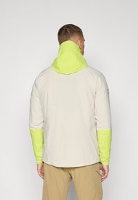 Jachetă ușoară dintr-un material bej, cu un glugă și accente pe mâneci de culoare verde intens. Prezintă un design simplu și o croială lejeră.